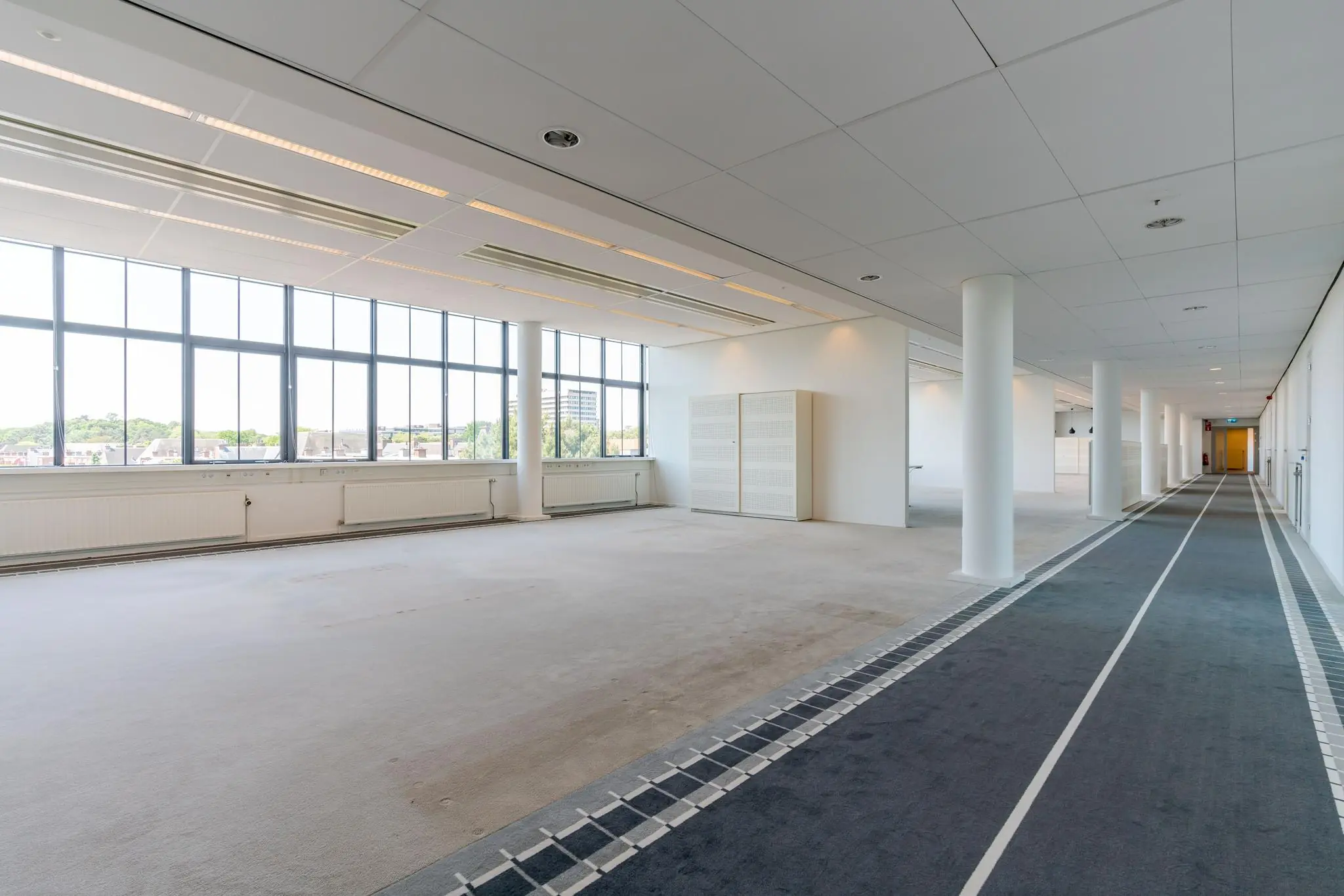 Lichte, lege kantoorruimte aan de Van Alkemadelaan met grote ramen en een brede gang met tapijtloper.